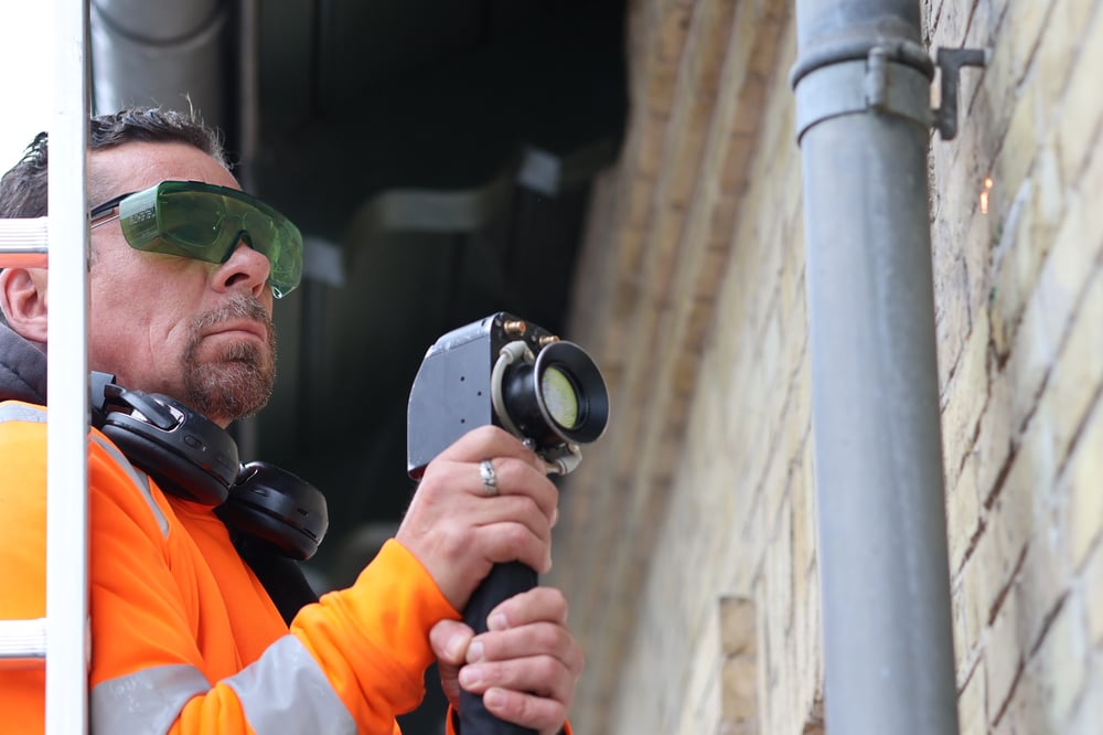 Håndværker med beskyttelsesbriller udfører kontrolleret laserrensning på en murstensfacade